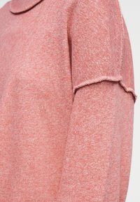 Μαλακό ροζ πλεκτό μανίκι πουλόβερ με λεπτομέρεια διπλής μανσέτας και διακριτή γραμμωτή υφή σε ανοιχτό φόντο.