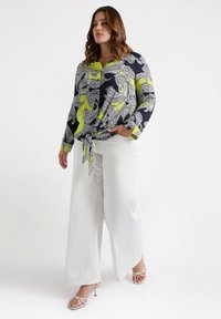 Blusa de patrón paisley en azul marino y verde lima con mangas largas, anudada a la cintura, combinada con pantalones anchos blancos y tacones con tiras.