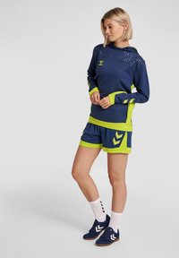 Completo sportivo blu navy con accenti verde neon; felpa con cappuccio presenta spalle texturate, abbinata a pantaloni corti coordinati e scarpe blu navy.