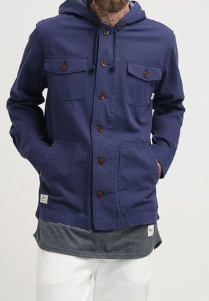 Mand iført en mørkeblå jakke med knapper og fire lommer, en hætte og en grå skjorte under, hænderne i jakkelommerne.