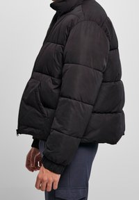Chaqueta de plumas negra con cuello alto, diseño acolchado y puños de canalé. Cuenta con cierre de cremallera y bolsillos laterales. Silueta estructurada y corta.