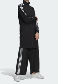 Černá tepláková souprava s dlouhou bundou na zip s vysokým límcem, doplněná o široké kalhoty, obojí s bílými pruhy.