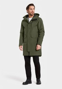 Grön vattentät rock med huva, dragkedja och tryckknappsstängning samt framfickor. Slät textur och något längre modell. Bärs över mörka byxor.