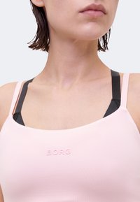 Κοντινό ενός ατόμου που φοράει ροζ αθλητικό σουτιέν με μαύρες λουρίδες ορατές από κάτω και τη λέξη «BORG» μπροστά.