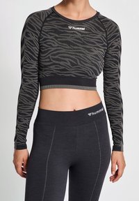 En person visar en figurnära, långärmad topp i svart och grått med ett vågmönster och matchande svarta leggings, vilket framhäver en aktiv livsstil.