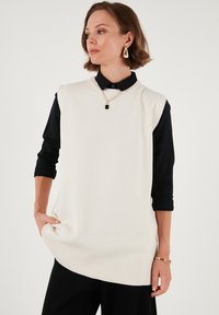 Knit sleeveless sweater in cream, featuring a ribbed hem. Worn over a black collared shirt, complemented by a gold chain necklace.