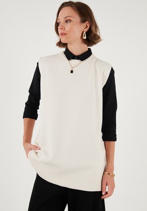 Knit sleeveless sweater in cream, featuring a ribbed hem. Worn over a black collared shirt, complemented by a gold chain necklace.