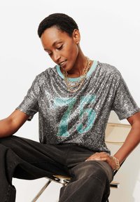 T-shirt à paillettes argentées avec un graphisme turquoise "75", manches courtes et col côtelé bleu clair, associé à un pantalon noir.