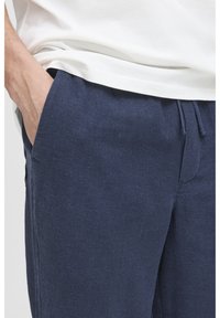 Pantaloni lejeri de culoare albastru închis, confecționați dintr-un material moale și texturat, cu talie elastică și buzunare laterale. Bluza este albă și ușor vizibilă.