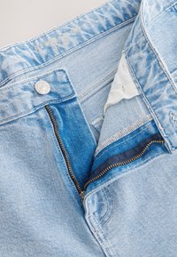Jean en denim bleu clair avec une braguette zippée, un bouton supérieur visible et un tissu intérieur en bleu plus foncé. Présente une couture simple et des bords effilochés.
