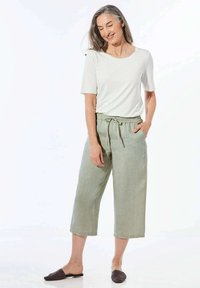 Weißes Kurzarmoberteil kombiniert mit hellgrünen Leinen-Culotte-Hosen mit elastischem Bund und seitlichen Taschen, ergänzt durch dunkle Slipper.