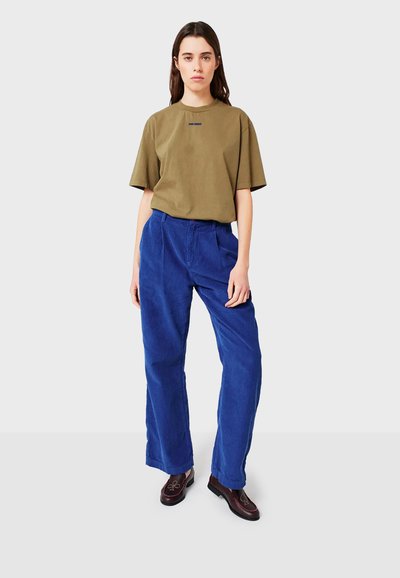 Donna con capelli castani lisci indossa una maglietta oversize verde oliva, pantaloni a gamba larga in corduroy blu brillante e mocassini marrone scuro con ricami.