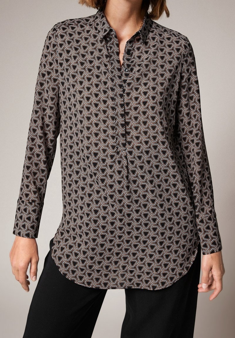 Femme portant une chemise noire à manches longues avec un motif géométrique hexagonal en blanc et beige, associée à un pantalon noir.