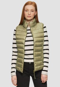 Helle olivgrüne Steppweste mit hohem Kragen, Reißverschluss und horizontalem Steppmuster, getragen über einem schwarz-weiß gestreiften Langarmshirt.