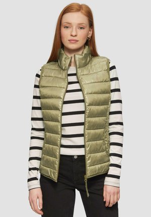 Gilet rembourré vert olive clair avec col haut, fermeture éclair et motif matelassé horizontal, porté sur une chemise à rayures noires et blanches à manches longues.