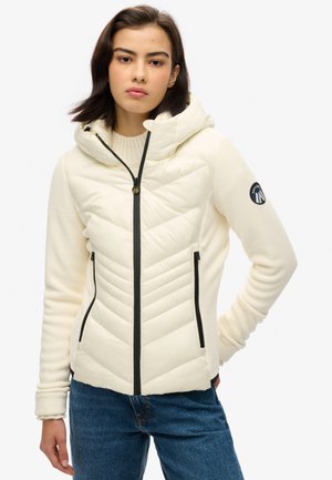 Giacca con cappuccio di colore bianco crema, caratterizzata da un davanti trapuntato con cuciture a zig-zag, tasche laterali con zip e un'etichetta con il logo sul maniche.