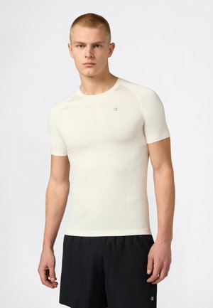 Modello maschile che indossa una maglietta atletica aderente bianco sporco a maniche corte e pantaloncini neri su uno sfondo bianco uniforme.