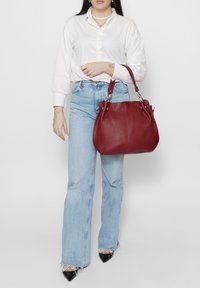 Borsa a tracolla in pelle rossa con forma arrotondata e Ferramenta in argento. Texture liscia con dettagli laterali. Indossata con jeans svasati azzurri chiari e camicia bianca.