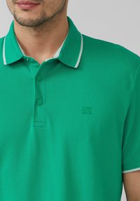 Zielona koszulka polo z kontrastowym jasnoniebieskim kołnierzykiem i wykończeniem, wyposażona w dwa guziki oraz wytłoczone logo na klatce piersiowej. Gładki, teksturowany materiał.