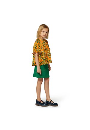 Helles gelbes Blumenbluse mit Rüschenschultern, kombiniert mit einem grünen Rock. Das Model trägt dunkelblaue Schuhe mit roten Akzenten und steht seitlich.