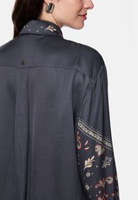 Dark, silky blouse featuring a patterned sleeve with floral and paisley designs in red and cream accents. Includes a prominent collar and button detail.