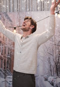 Ljusgrå cardigan knäppt fram, bärs över en vit skjorta. Bakgrunden visar en snöig gatumiljö med festlig belysning.
