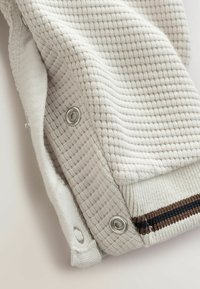 Pull en beige clair avec texture, présentant des détails en tricot gaufré, doté de boutons-pression argentés et d'une bordure rayée en côtes marron et bleu marine.