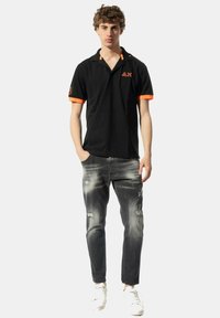 Polo nero con dettagli arancioni, caratterizzato da un colletto e maniche corte. Indossato con jeans grigi strappati e sneaker bianche.