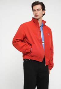 Κόκκινο bomber μπουφάν με ψηλό γιακά, ελαστικούς μανσέτες και εφαρμοστή μέση. Φοριέται πάνω από ένα μπλε πουκάμισο, που διαθέτει μια διακριτική λεπτομέρεια λογότυπου.