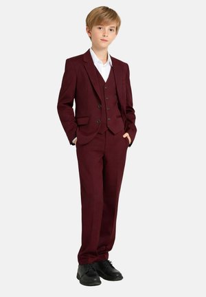 Garçon debout les mains dans les poches, portant un costume trois pièces bordeaux, une chemise blanche et des chaussures habillées noires sur un fond uni.