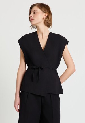 Frau mit kurzen Haaren trägt ein ärmelloses schwarzes Wickeloberteil, das an der Taille gebunden ist, kombiniert mit schwarzen weit geschnittenen Hosen vor neutralem Hintergrund.