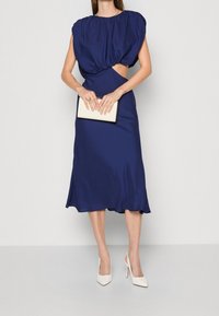 Marinblå midiklänning med rynkad halsringning, vriden frontdesign, sidoutskärning och svävande nederdel. Matchad med en vit clutch och klackar.