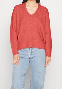 Röd v-ringad tröja med ribbat mönster och lös passform, kombinerad med ljusblå jeans. Har långa ärmar och sidslitsar.