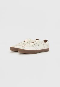 Zapatos de lona crema con suelas de goma marrón, que presentan un diseño de cordones y acentos marrones contrastantes en la puntera y los lados.
