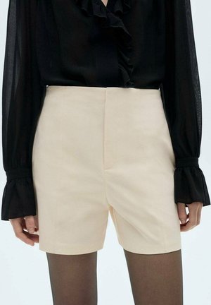 Sort, langærmet, gennemsigtig bluse med flæseærmer og halsudskæring, kombineret med højtaljede cremefarvede shorts og mørke gennemsigtige strømpebukser.