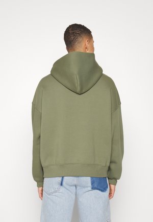 Homme portant un sweat à capuche vert olive et un jean bleu clair avec un patch bleu foncé, debout de dos sur un fond blanc uni.