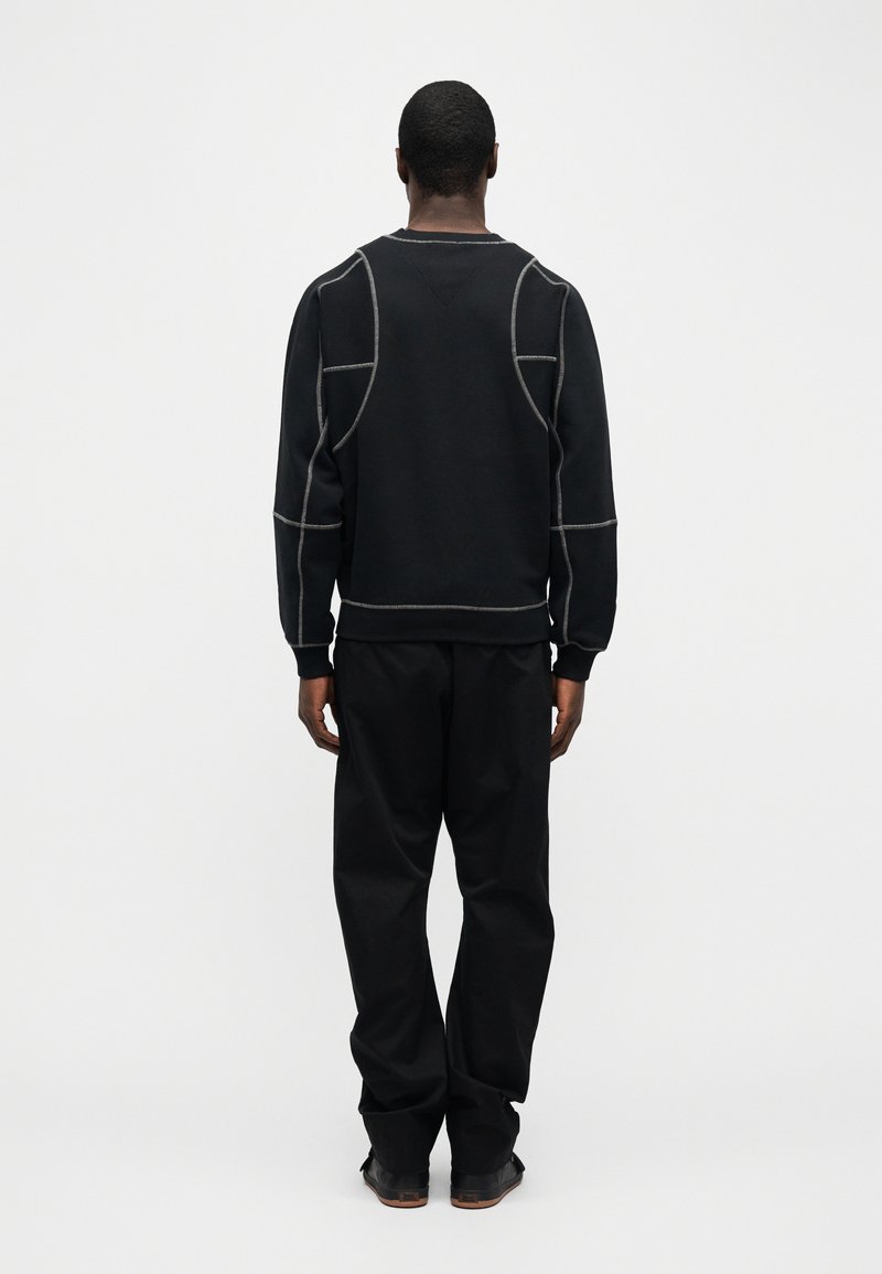 Uomo in piedi di spalle, indossa pantaloni neri e una felpa nera con dettagli di cuciture bianche a contrasto, su uno sfondo bianco semplice.