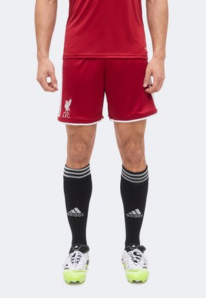 Männlicher Athlet trägt rote Liverpool FC-Shorts, schwarze Adidas-Kniestrümpfe und weiße Fußballschuhe mit neongrünen Sohlen, steht vor einfarbigem Hintergrund.