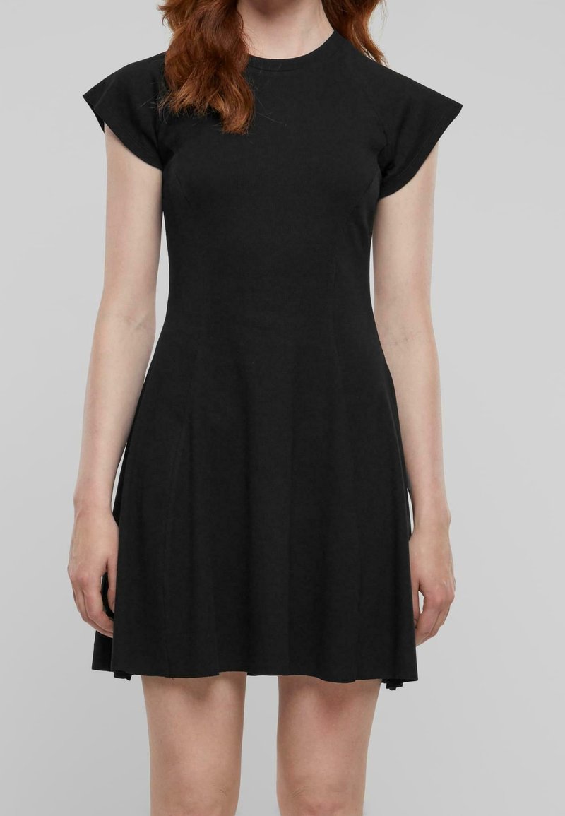 Femme portant une robe noire courte avec des manches courtes et une taille cintrée, debout devant un fond gris uni.