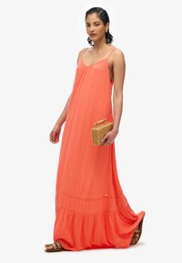 Długa koralowa sukienka na ramiączkach spaghetti, z lekko opadającym dolnym tierem, teksturowana tkanina oraz okrągła torebka ze słomy. Noszona z brązowymi sandałami na płaskiej podeszwie.