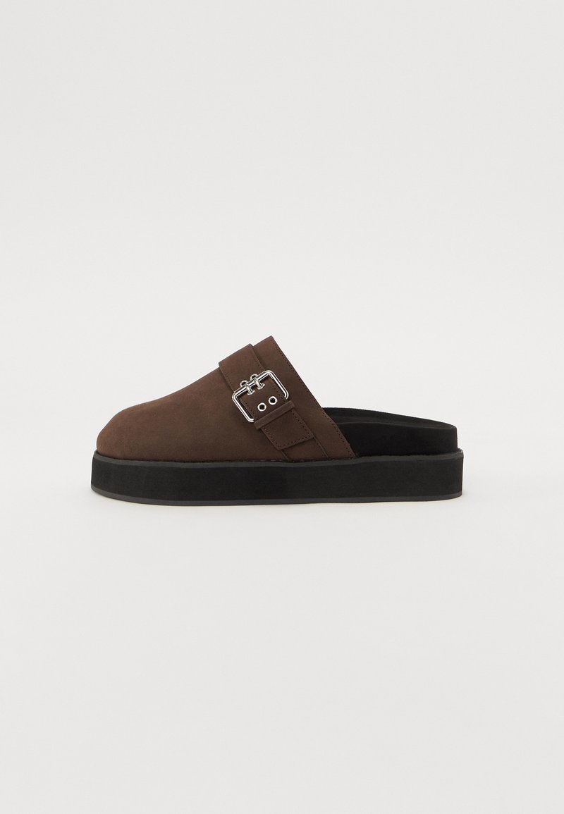 Brown suede slip-on shoes with a rounded toe, black sole, and a silver buckle detail on the strap. Flat and textured design.
