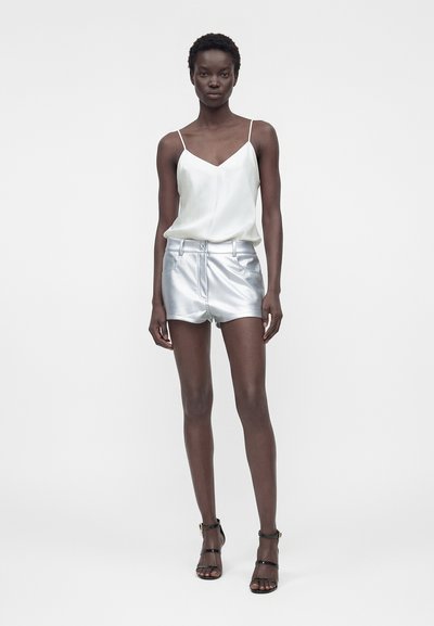 Femme portant un caraco en satin blanc, un short métallisé argent, et des sandales noires à talons hauts avec des lanières, debout devant un fond uni.