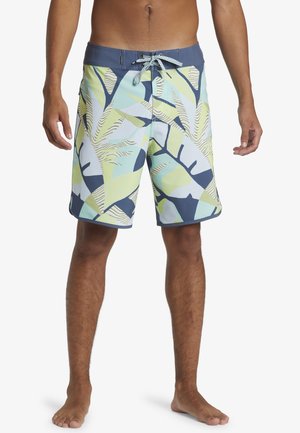 Hombre con bañador de longitud a la rodilla con patrones de hojas tropicales en azul, verde y beige, y cintura con cordón azul, de pie descalzo.