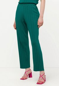 Pantalones plisados en color verde azulado con cintura alta y corte recto. Combinados con tacones rosas de tiras, que presentan un tacón ancho.