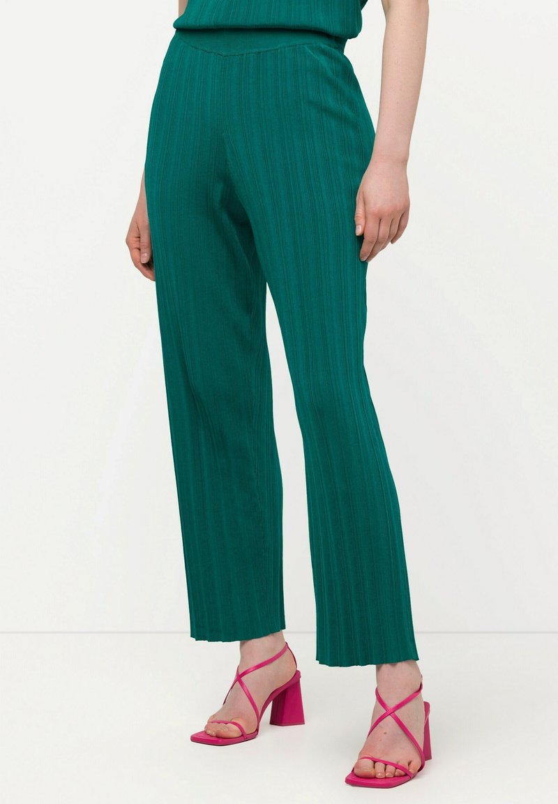 Pantalones plisados en color verde azulado con cintura alta y corte recto. Combinados con tacones rosas de tiras, que presentan un tacón ancho.
