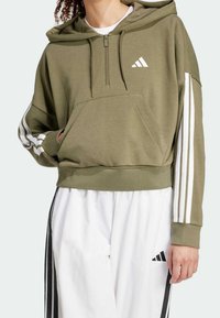 Olivgrön kort hoodie i mjukt material, med dragkedja framtill, en kenguruficka och vita tre-stripes detaljer på ärmarna.