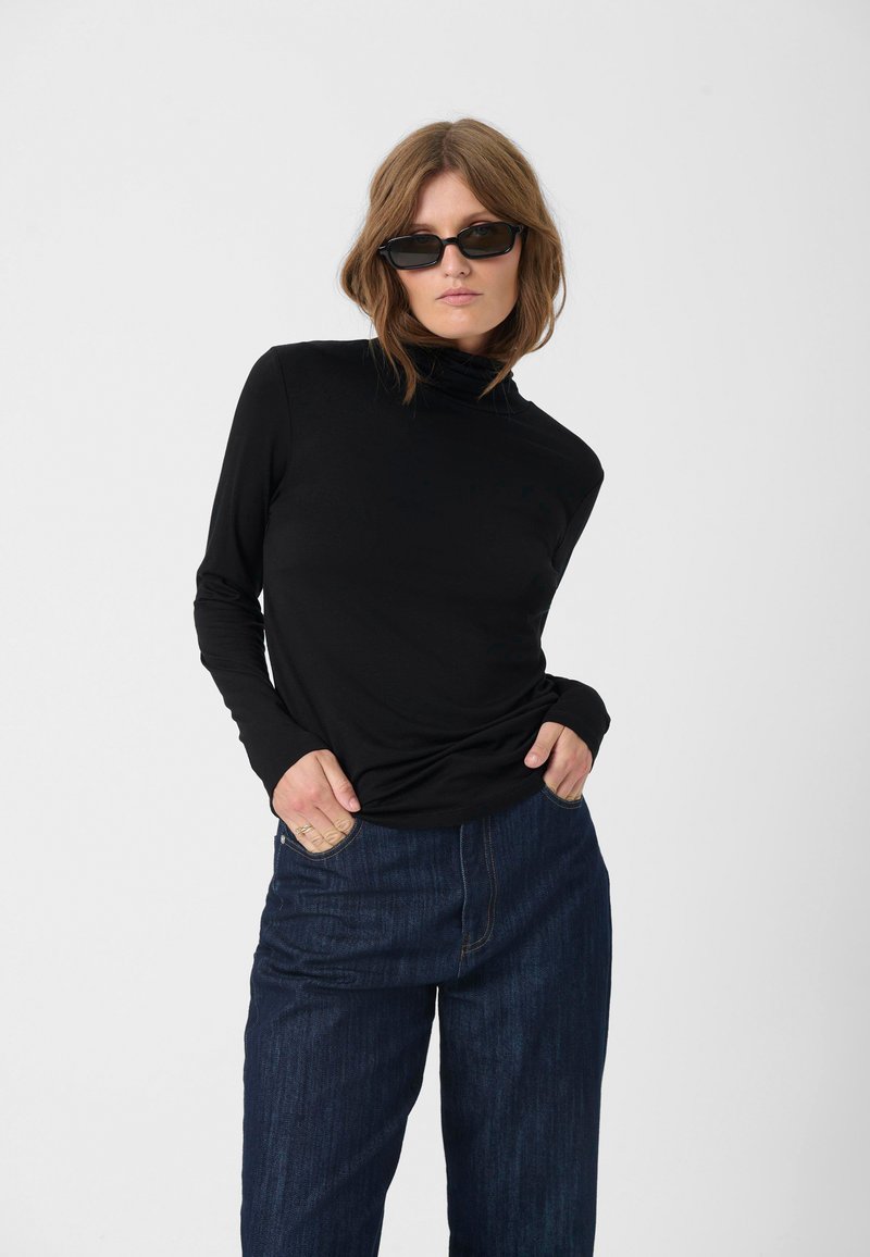 Mustast pikkade varrukatega kõrge kaelusega pluus, mis on kokku sobitatud siniste laiade jalgade teksadega. Pluus on valmistatud siledast kangast; teksad näitavad veidi tekstureeritud viimistlust.