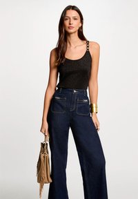 Débardeur noir ajusté avec texture pailletée et bretelle en chaîne. Jean en denim foncé taille haute, à jambe large, avec des coutures contrastantes et des poches. Sac à main marron avec franges.
