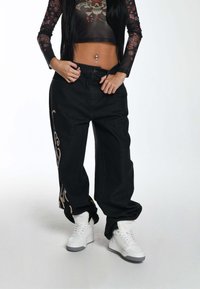 Pantalones baggy de mezclilla negra con un patrón decorativo beige a lo largo del costado, combinados con zapatillas altas blancas y una blusa larga de manga larga de flores transparente.