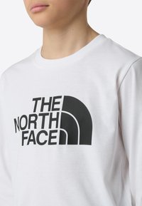 Fehér, hosszú ujjú pamut póló, elöl jól látható, fekete „THE NORTH FACE” logóval.